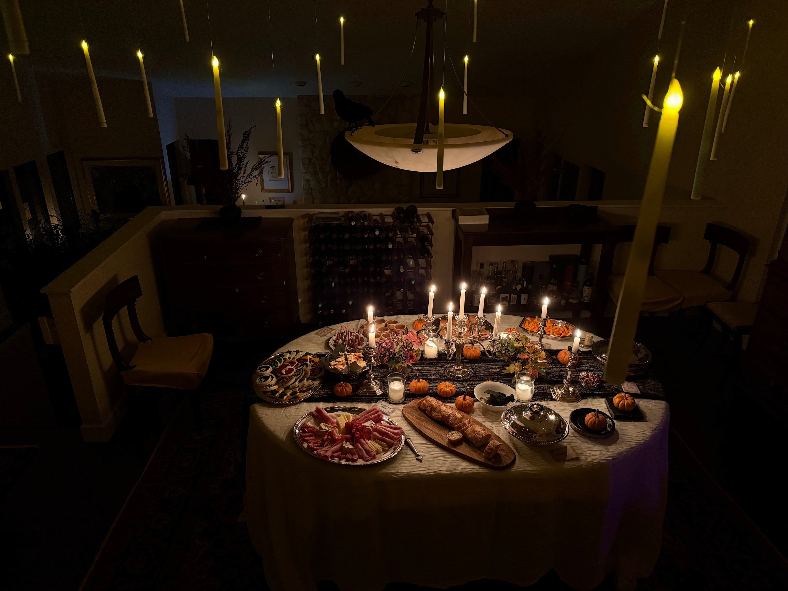 A dimly lit dining table set with candles, bread, sliced meats, and small dishes in a dark room with hanging candles and a pendant light.