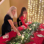 Two women decorating a table with a red tablecloth, greenery, and candles against a string light backdrop.