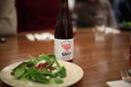 A bottle of Uber wine with a white label stands on a wooden table next to a plate of green salad, with blurred people in the background.