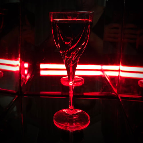 A clear stemmed wine glass with a wide bowl, shown on a dark reflective surface with red neon lighting and mirror reflections in the background.