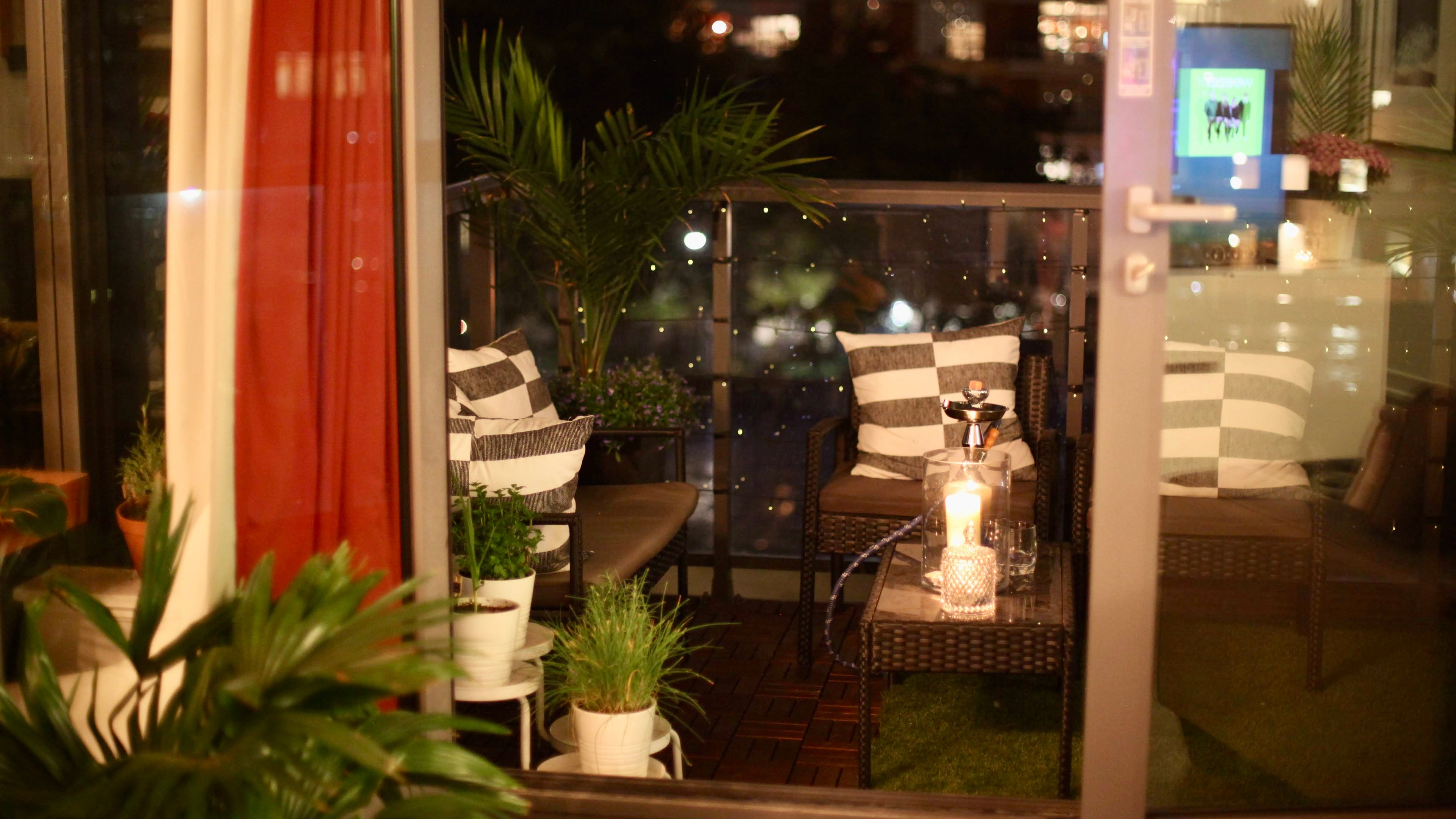 A cozy outdoor patio seating area viewed through a glass door at night, featuring striped cushions, potted plants, string lights, and a small table with a candle.