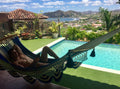 A man relaxes in a blue and beige patterned hammock strung over a turquoise swimming pool, with a scenic ocean and mountain view in the background.