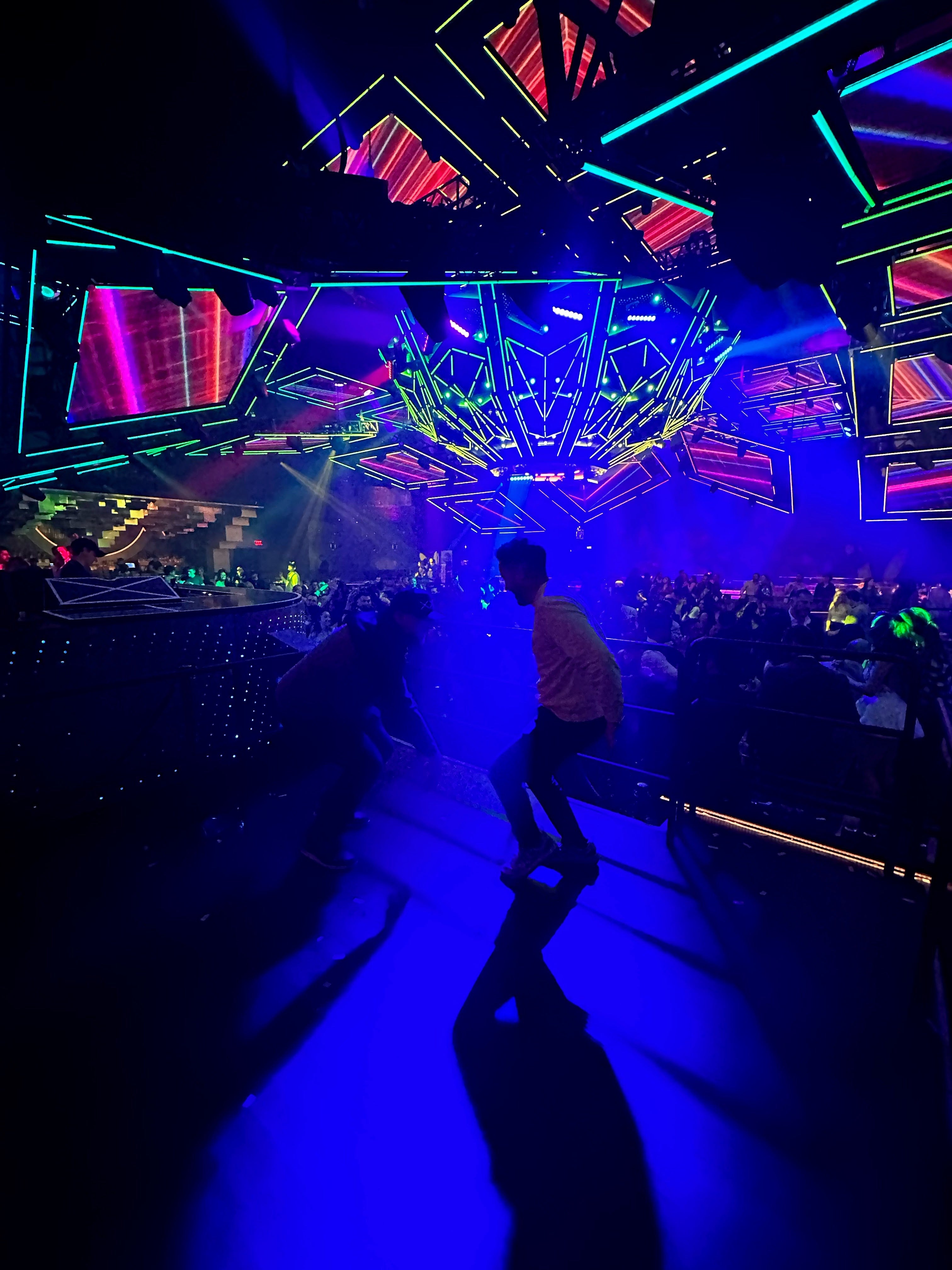 A person stands on a brightly lit stage or runway under dramatic blue, purple, and pink neon lighting, with a dark venue interior and audience visible in the background.