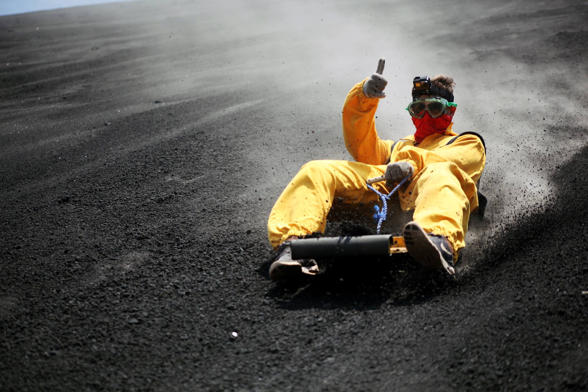 A person wearing a bright yellow hazmat-style suit, red face covering, and goggles slides on a dark volcanic or gravel surface, with a cloud of dust behind them.