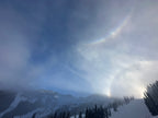 A snowy mountain landscape with evergreen trees in the foreground and a faint rainbow arching through the cloudy sky.
