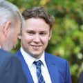 Man in a blue suit and tie standing outdoors with greenery in the background