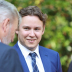 Man in a blue suit and tie standing outdoors with greenery in the background
