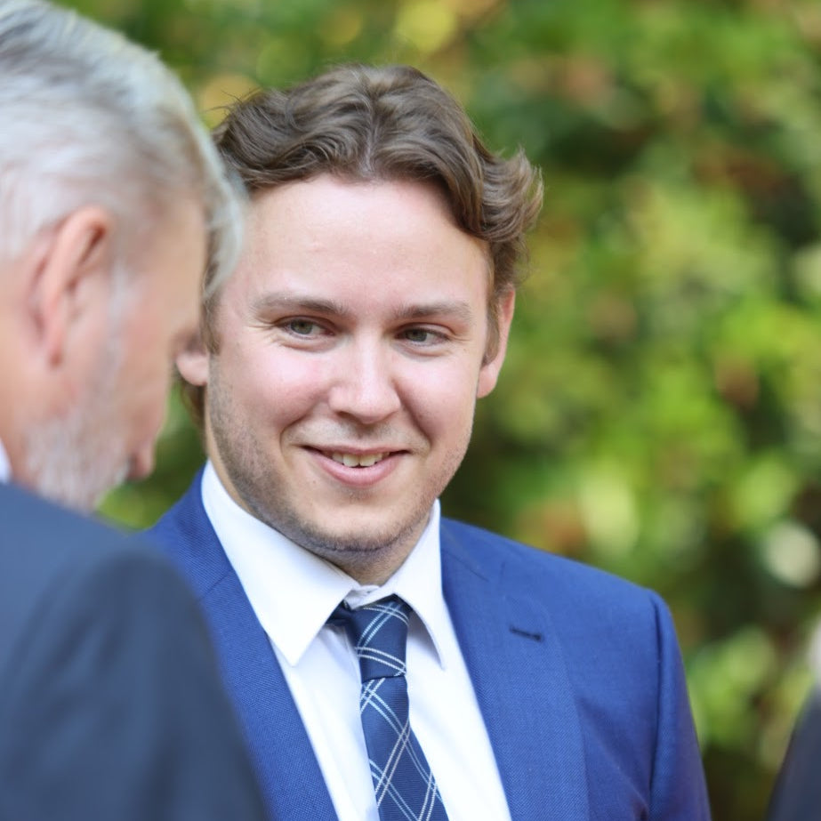 Man in a blue suit and tie standing outdoors with greenery in the background