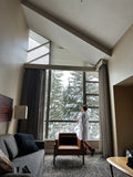 Living room with large windows overlooking trees, featuring a gray sofa and a person standing.