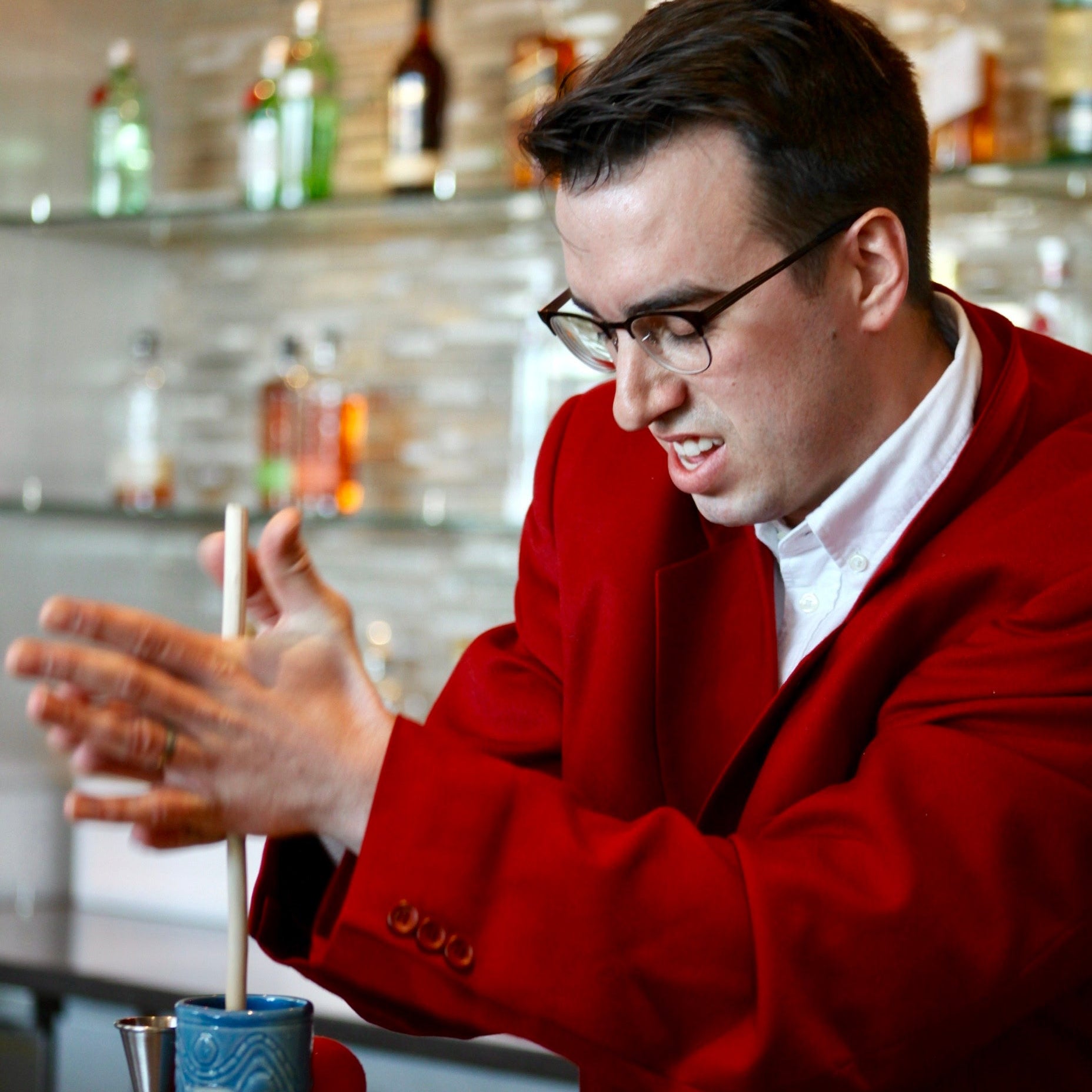 Man in a red jacket mixing a drink at a bar with bottles in the background