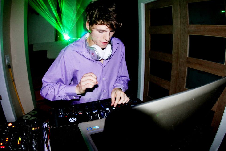 A man wearing a purple button-up shirt and white over-ear headphones is DJing at a turntable, with a laptop in the foreground and green laser lights in the background.