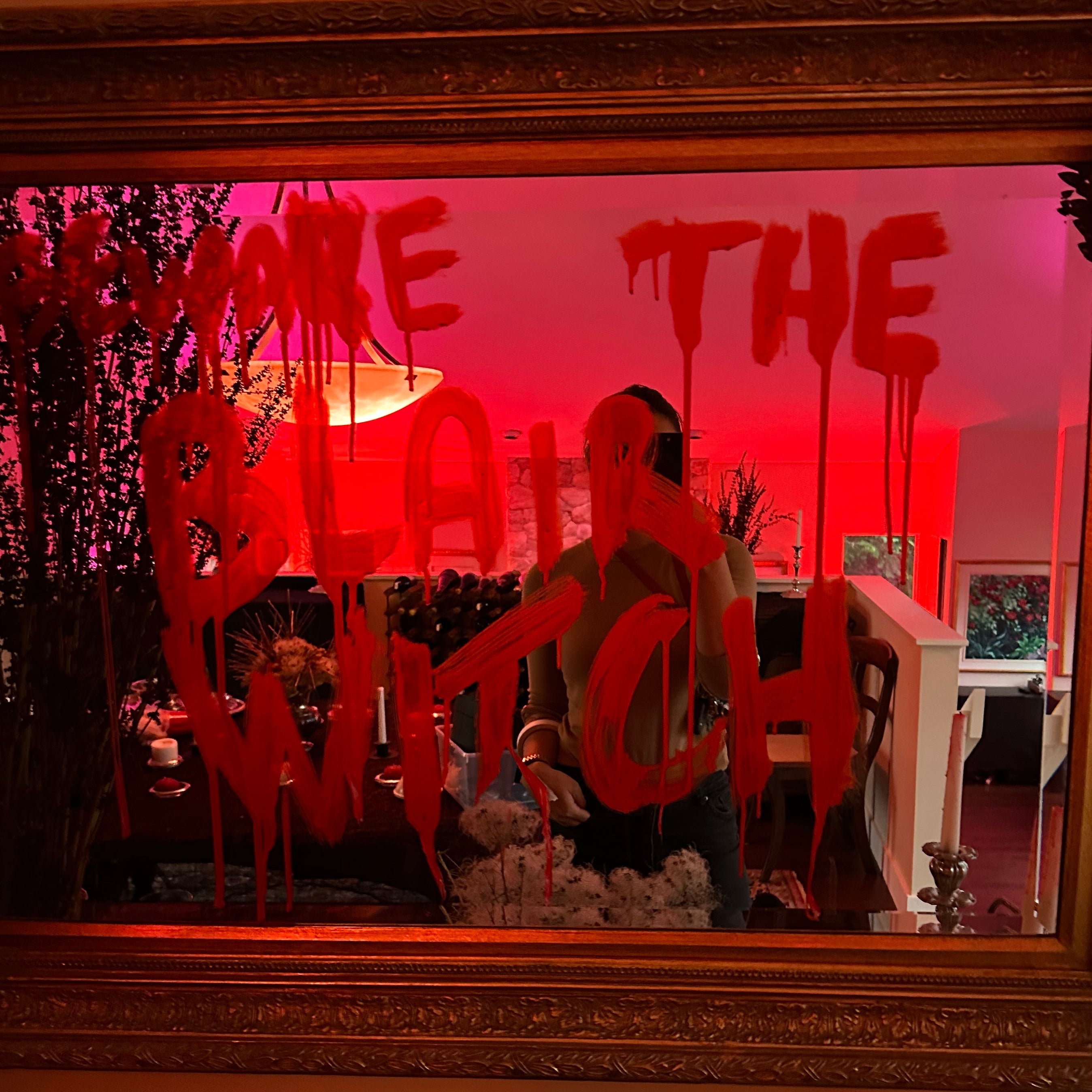 A large ornate gold-framed mirror on a wall, reflecting a person in a dark outfit while the glass is covered in dripping red spray-paint text that reads 'beware the blair WITCH'. The scene is lit with strong red ambient light, and a candle holder is visible in the foreground.