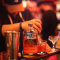 Bottle of Bulleit Bourbon on a bar with a bartender's hand and other drinks in the background.