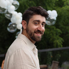 Man with a beard and mustache standing outdoors with white balloons and greenery in the background