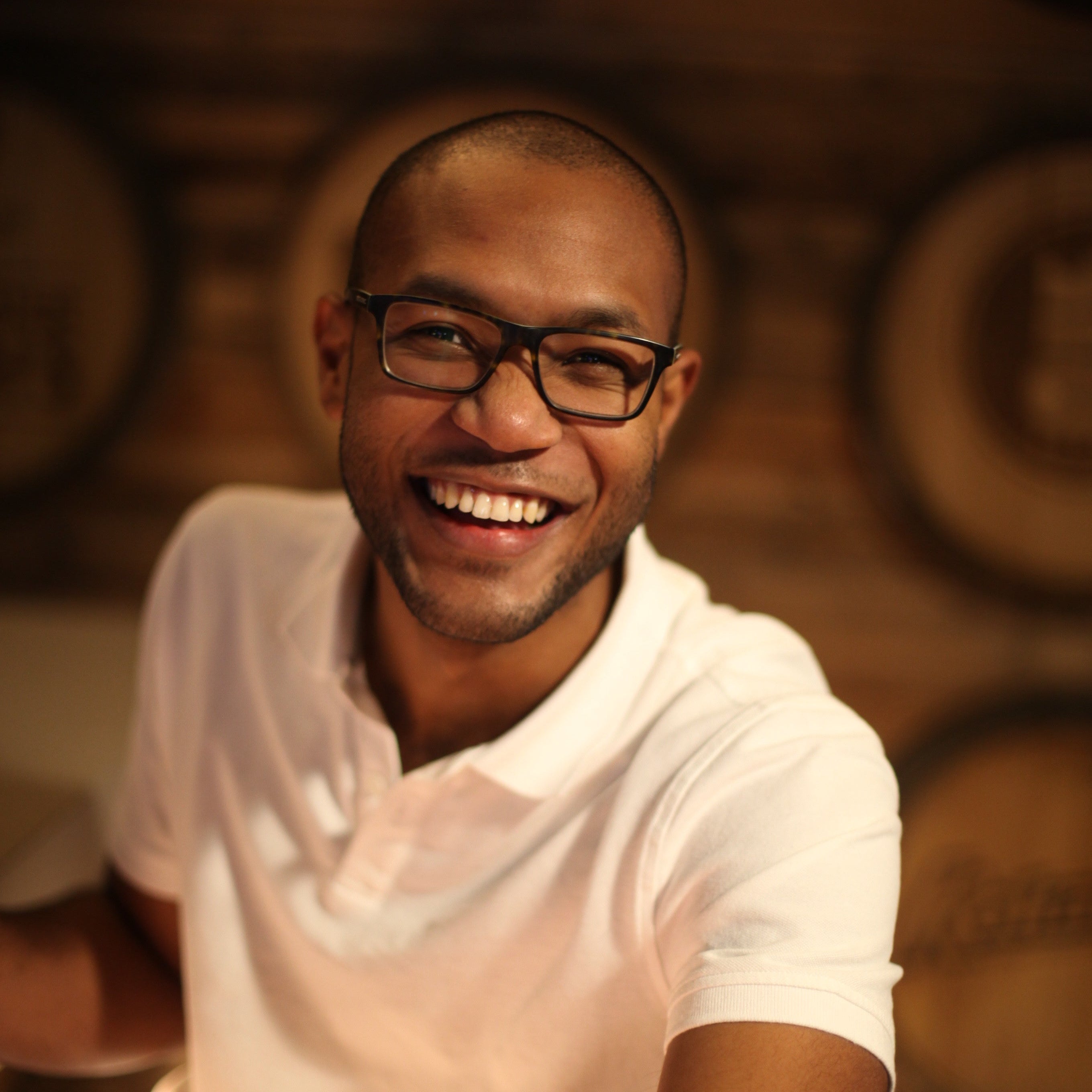 Man wearing glasses and a white shirt smiling in front of wooden barrels.