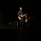 David Martin and another musicians on stage with guitars and a microphone, performing in front of a dark background.