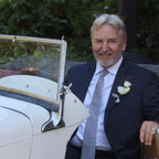 David Martin in a suit sitting in an old-fashioned MG car with a white flower boutonniere.