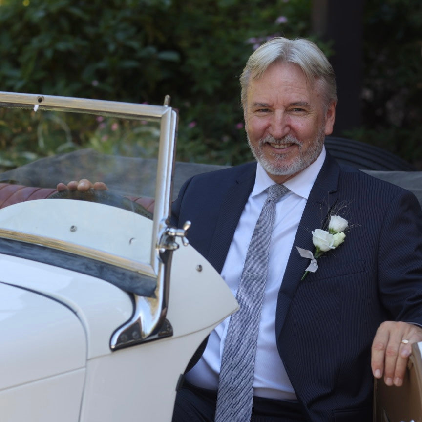 David Martin in a suit sitting in an old-fashioned MG car with a white flower boutonniere.