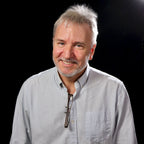 David Martin with gray hair and beard wearing a light blue shirt against a black background