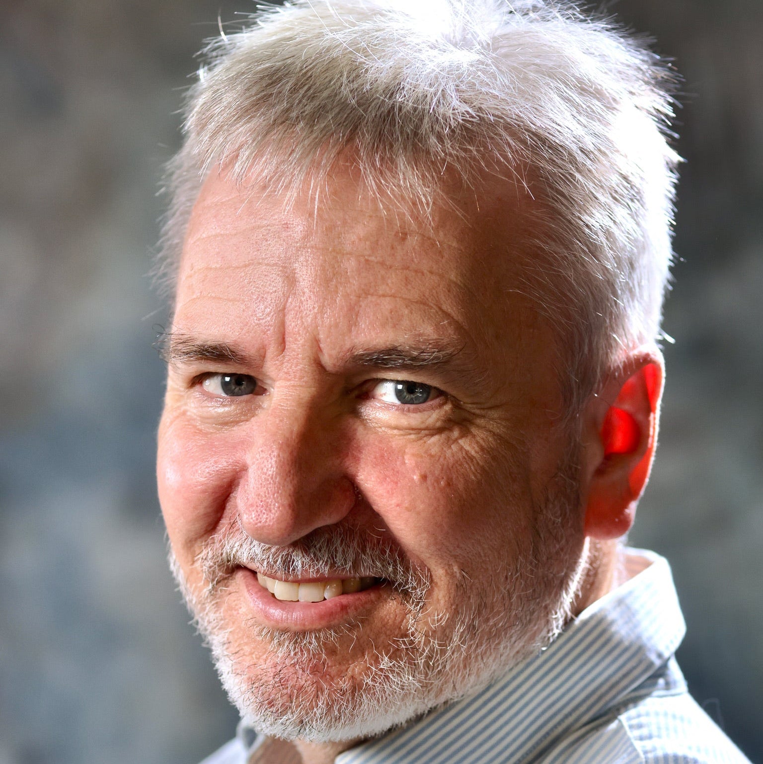 Man with gray hair and beard wearing a light-colored shirt against a blurred natural background