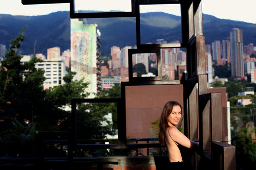 Evelyn Martin with long brown hair stands on a balcony framed by dark geometric wooden structures of a luxury hotel at a luxury resort, looking over the Colombian city skyline with mountains in the background.