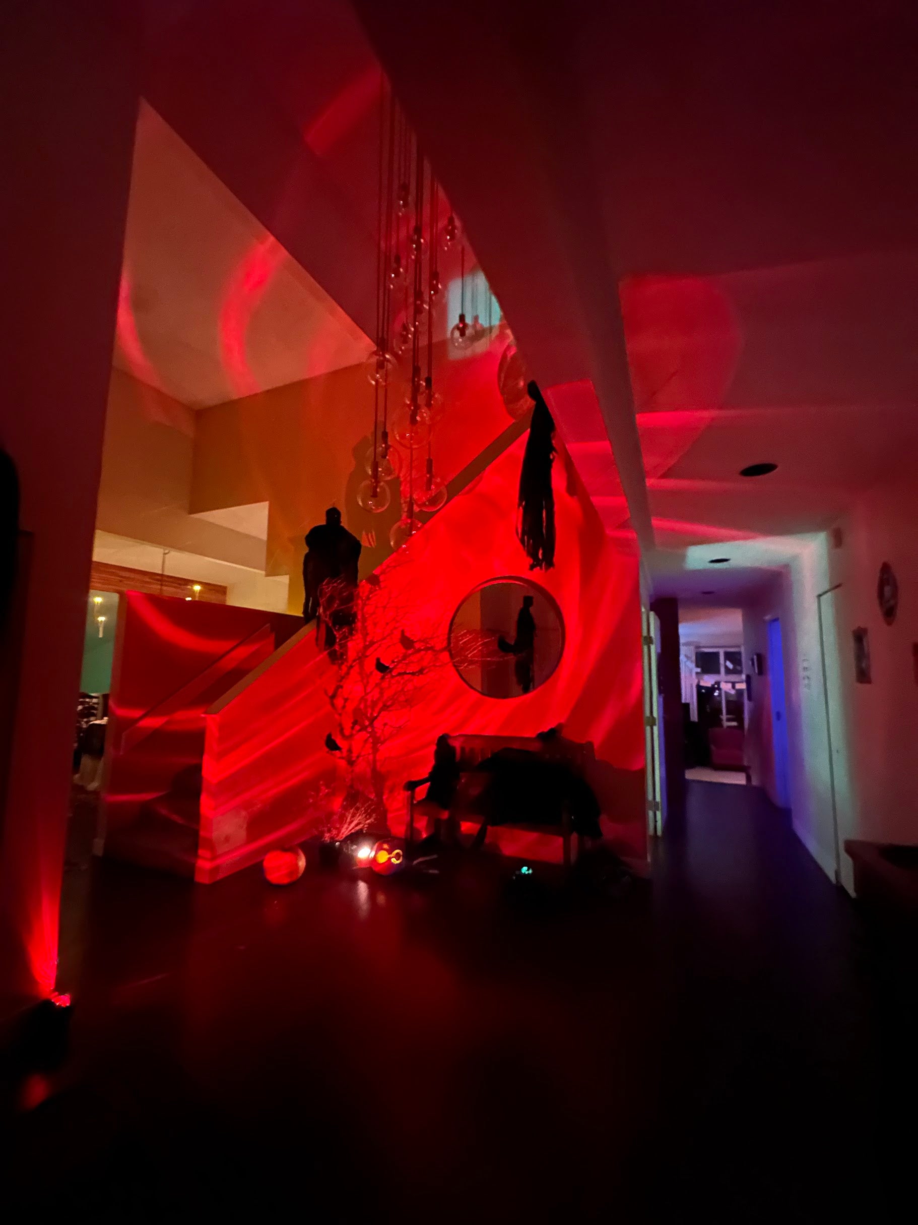 A dimly lit interior hallway and staircase area illuminated by strong red lighting, with hanging decorative lights and a circular wall mirror visible. No clear sellable product is isolated; this reads more like an atmospheric room scene or environment shot.