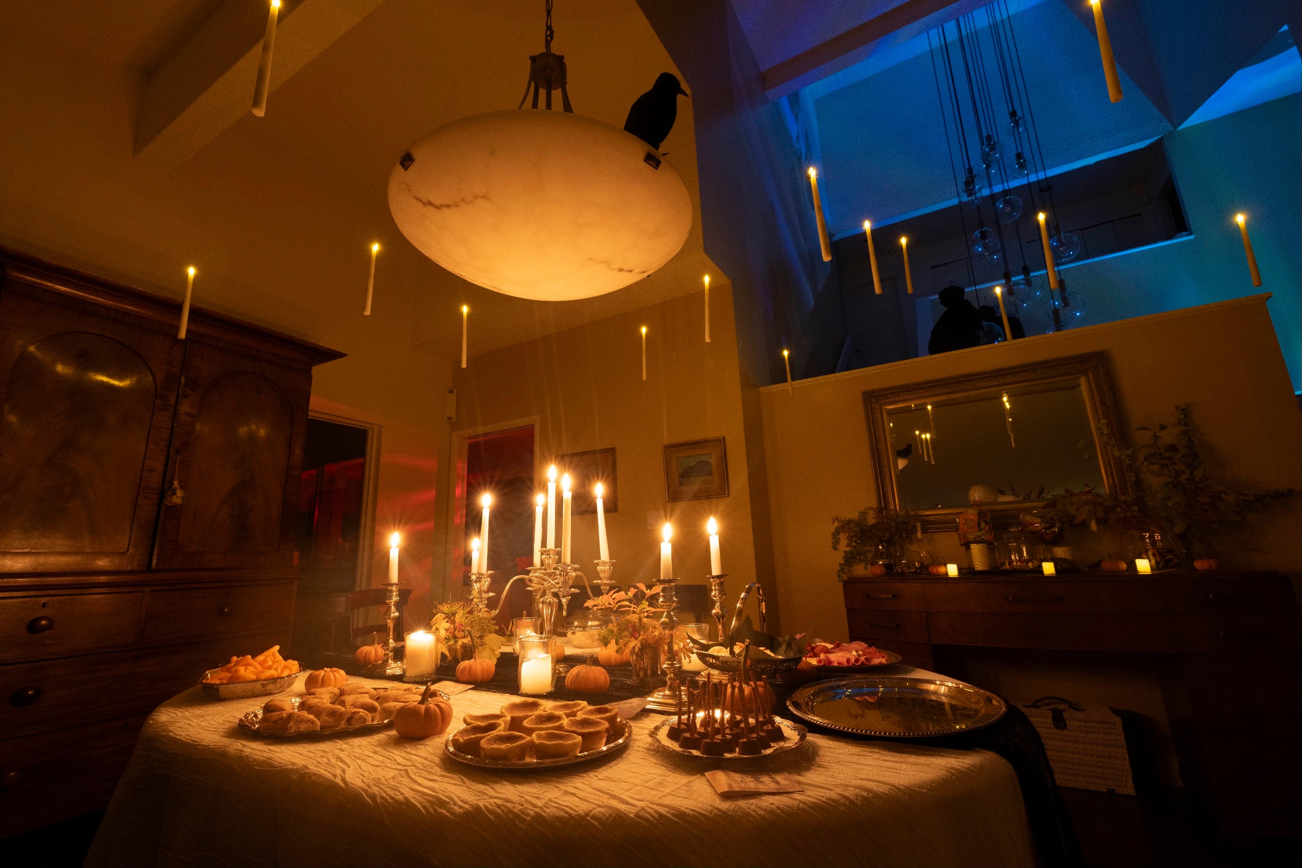 A dimly lit dining room with a table covered in candles, food, and decorative objects, under a large hanging lamp and blue ambient lighting in the background.