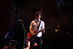 A male musician performing on stage, holding a red electric guitar and wearing a white shirt with a patterned tie against a dark stage background.