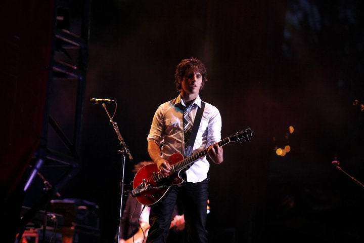 A male musician performing on stage, holding a red electric guitar and wearing a white shirt with a patterned tie against a dark stage background.