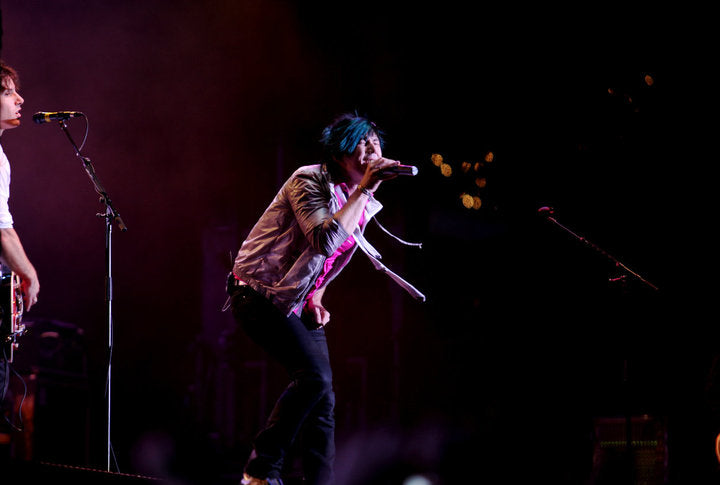 A live concert performer singing into a microphone on stage, wearing a patterned shirt and dark pants, with stage lighting and a dark background.