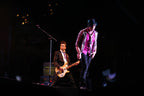 A live concert photo of a male musician performing on stage, wearing a pink shirt, a shiny silver jacket, and dark pants, with another guitarist and a microphone visible in the background under dramatic stage lighting.