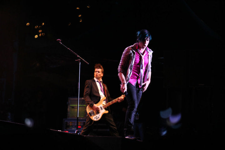 A live concert photo of a male musician performing on stage, wearing a pink shirt, a shiny silver jacket, and dark pants, with another guitarist and a microphone visible in the background under dramatic stage lighting.