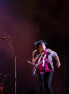 A live concert photo of a performer singing into a microphone on stage, wearing a pink top, a light-colored jacket, and dark pants under dramatic stage lighting.
