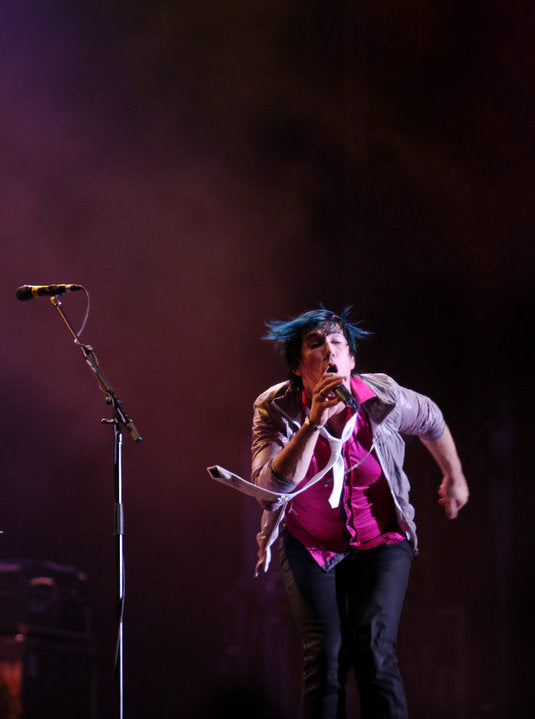 A live concert photo of a performer singing into a microphone on stage, wearing a pink top, a light-colored jacket, and dark pants under dramatic stage lighting.