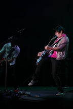 A live concert photo of a musician performing on stage, playing a blue and white electric guitar while wearing a pink shirt, a metallic silver jacket, and black pants. Another band member is partially visible in the background under dim stage lighting.