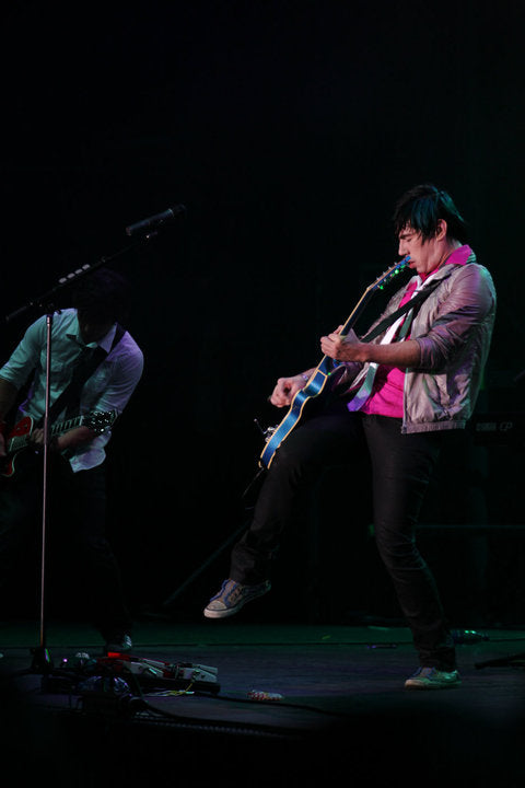 A live concert photo of a musician performing on stage, playing a blue and white electric guitar while wearing a pink shirt, a metallic silver jacket, and black pants. Another band member is partially visible in the background under dim stage lighting.