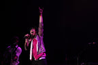 A live concert photo of a singer performing on stage, holding a microphone and raising one hand in a rock-and-roll gesture. The background is dark with stage lighting casting a pinkish-purple glow on the performer.