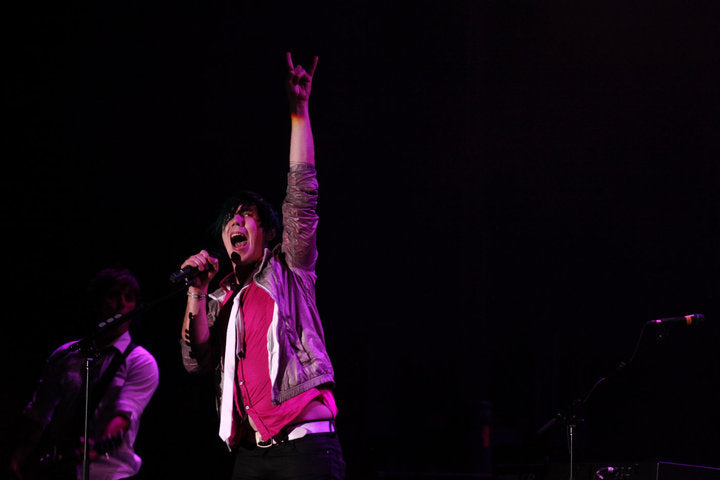 A live concert photo of a singer performing on stage, holding a microphone and raising one hand in a rock-and-roll gesture. The background is dark with stage lighting casting a pinkish-purple glow on the performer.