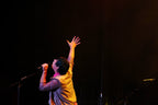 A male singer performing on stage with one arm raised, holding a microphone. He is wearing a yellow shirt and a pink bandana, set against a dark stage background with stage lighting.