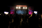 A nighttime concert scene with a stage lit by bright overhead lights, large side screens, and a crowd of people standing in the foreground. A red sign above the stage reads 'THE CONCERT STAGE 100'.
