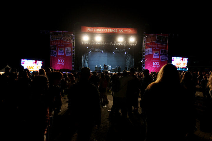 A nighttime concert scene with a stage lit by bright overhead lights, large side screens, and a crowd of people standing in the foreground. A red sign above the stage reads 'THE CONCERT STAGE 100'.
