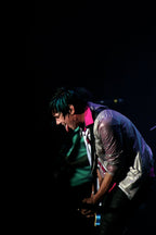 A person in a shiny silver jacket and bright pink shirt performing on stage against a dark background.