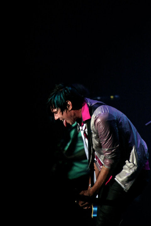A person in a shiny silver jacket and bright pink shirt performing on stage against a dark background.