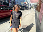 A woman walking on a city sidewalk wearing a black oversized t-shirt dress with a yellow outlined graphic on the front. A red car is parked on the left, and buildings line the street in the background.