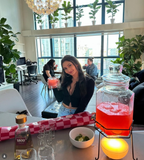 A woman in a black crop top and light blue jeans sits in a bright, modern lounge bar holding a pink drink. In the foreground, a glass pitcher filled with pink liquid sits on a black wire stand next to a bowl of lime wedges and shot glasses.