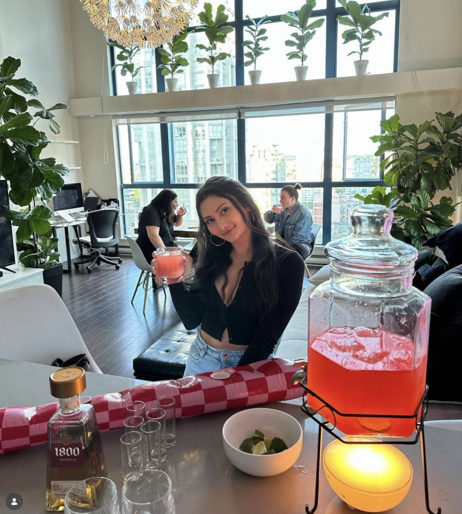 A woman in a black crop top and light blue jeans sits in a bright, modern lounge bar holding a pink drink. In the foreground, a glass pitcher filled with pink liquid sits on a black wire stand next to a bowl of lime wedges and shot glasses.