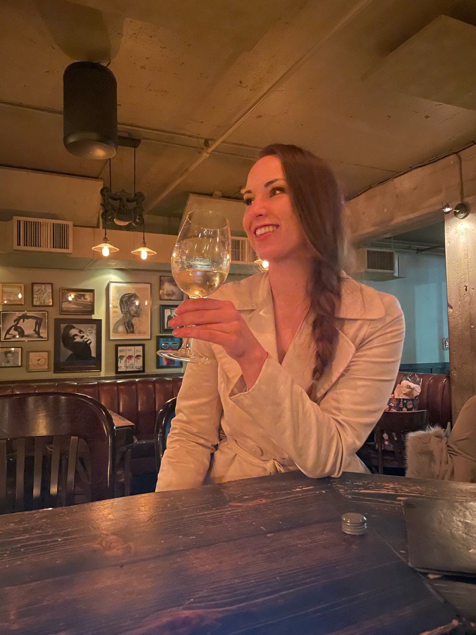 Evelyn Martin at parlour bar in Vancouver with braided hair and a beige trench coat holding a glass of wine in a cozy bar setting.
