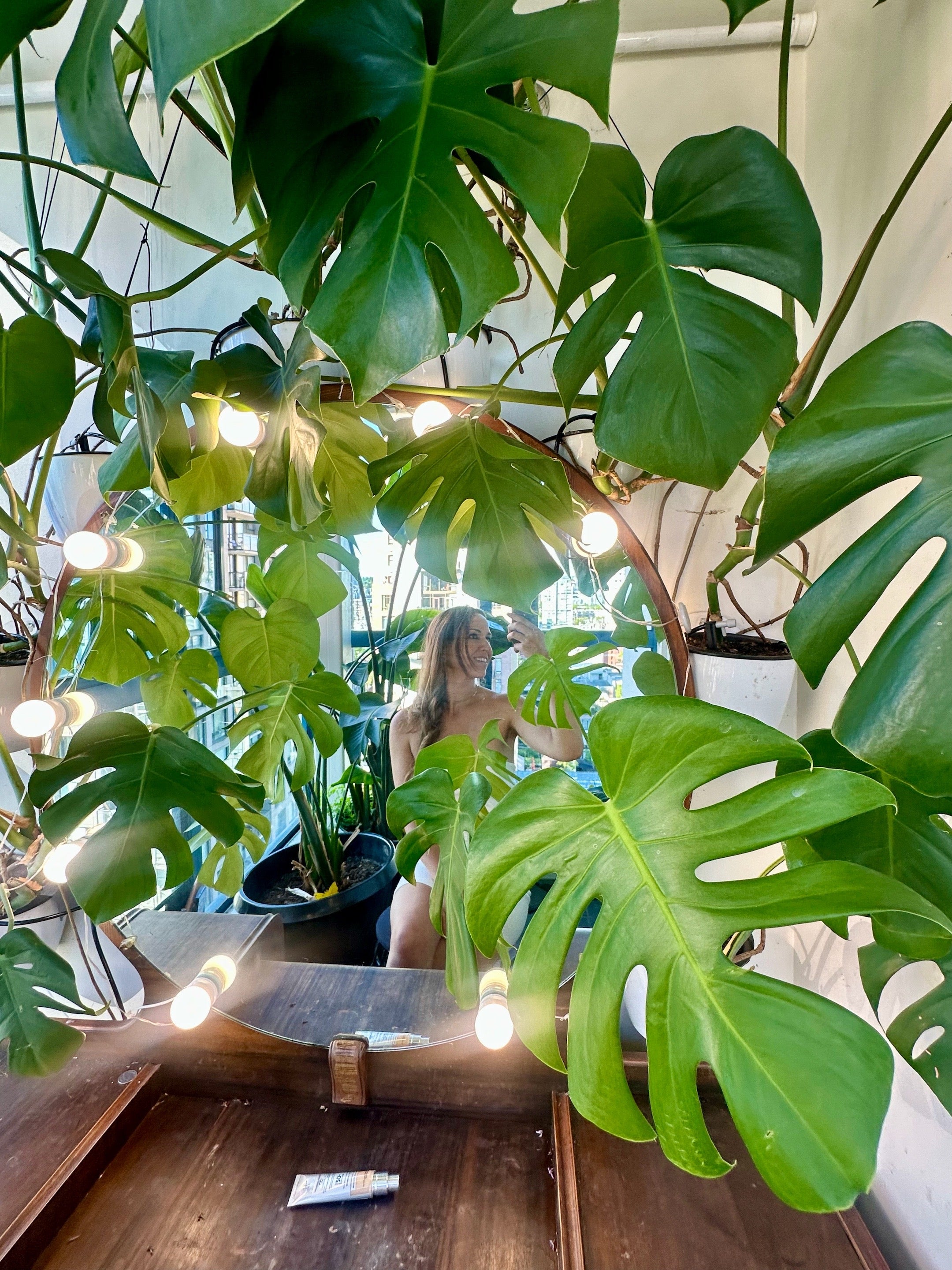 Evelyn Martin sitting among lush green plants in her Greenroom beauty makeup room with it cosmetics on the counter