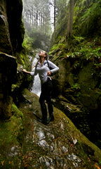 Evelyn Martin standing in a forest with a waterfall in the background doing a photoshoot. 