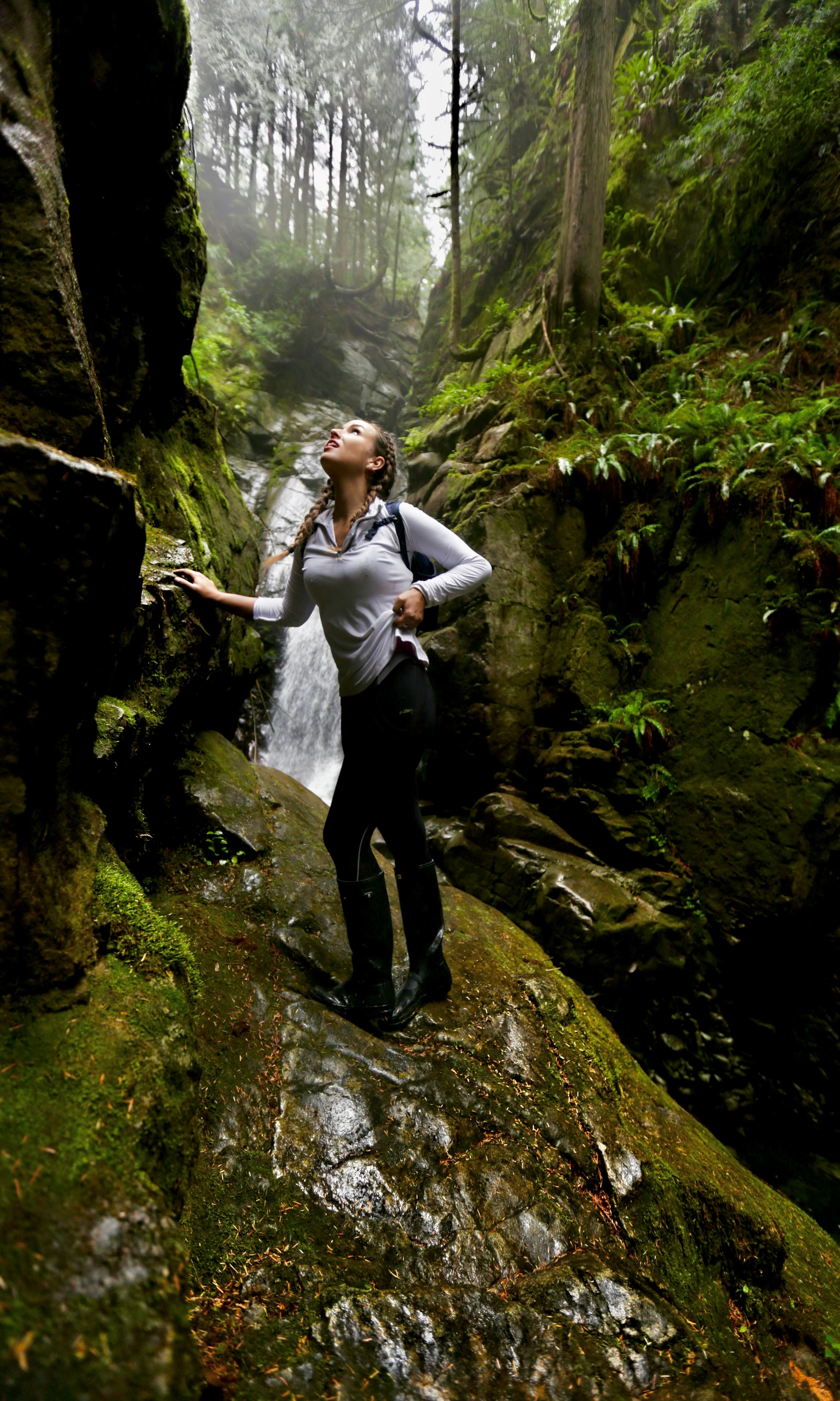 Evelyn Martin standing in a forest with a waterfall in the background doing a photoshoot. 